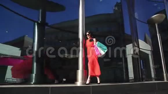 美丽的年轻女孩穿着红色连衣裙手里拿着购物袋享受着美丽的天气女孩沿着视频的预览图