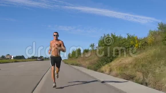 年轻的微笑肌肉男戴着太阳镜在高速公路上慢跑用他的智能手机耳机听音乐男性视频的预览图