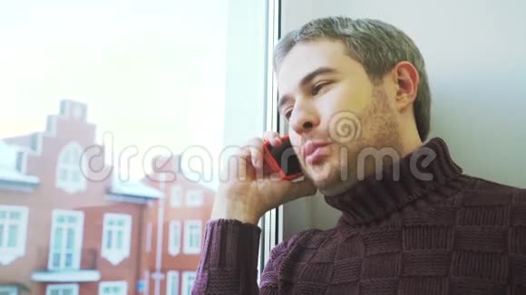 一个长着短胡子的开朗的年轻人靠窗用手机说话视频的预览图