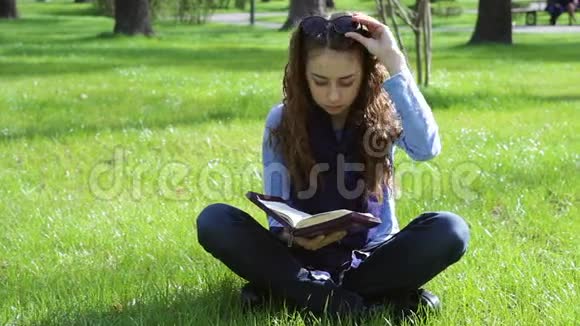 美丽的年轻女子坐在草地上的一个夏天的公园里读一本圣经视频的预览图