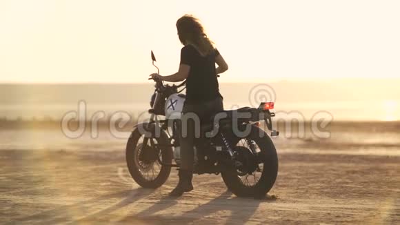 有魅力的年轻女摩托车手在摩托车上精疲力竭然后骑走日落或日出射向沙漠女自行车手视频的预览图