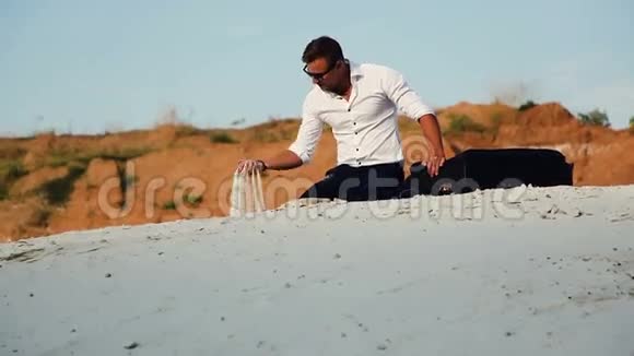 一个穿着商务服拿着行李包坐在沙滩上的年轻人在手指间撒满沙子视频的预览图