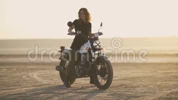 有魅力的年轻女摩托车手在摩托车上精疲力竭然后骑走日落或日出射向沙漠女自行车手视频的预览图