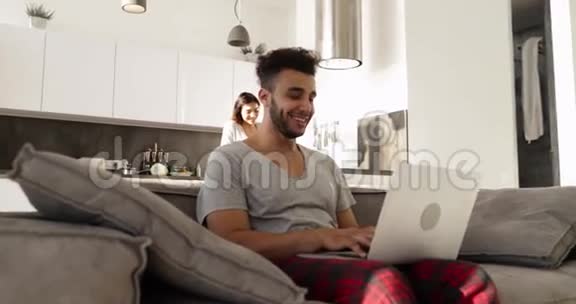 快乐年轻混合种族夫妇家庭内部西班牙男子使用笔记本电脑亚洲妇女烹饪早餐视频的预览图