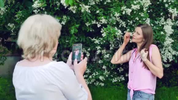 年长的女人给年轻漂亮的女孩拍照妈妈给女儿拍照背景中的白色花朵视频的预览图