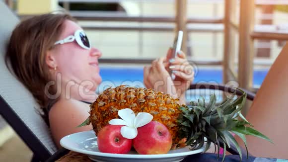 夏天戴着太阳镜躺在日光浴床上的年轻女性使用智能手机在游泳池旁边发送信息视频的预览图