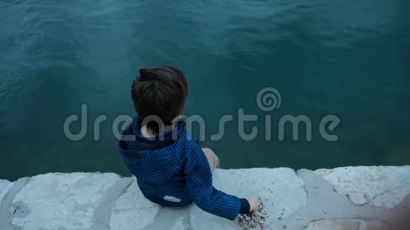坐在码头上的小男孩把石头扔到户外的水里视频的预览图