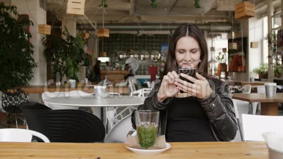 时尚的年轻女士喝着草药茶用智能手机坐在现代咖啡馆的桌子上微笑着4K视频的预览图