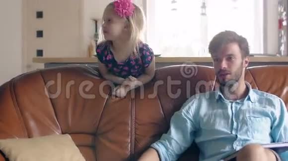 年轻的父亲坐在沙发上和别人说话而他的女儿在后面微笑4k视频的预览图