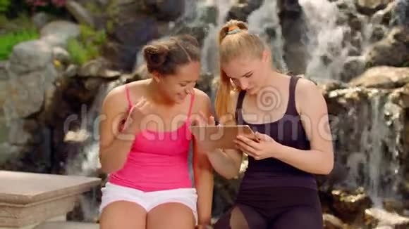 女孩们在户外平板电脑上看照片年轻女子坐在瀑布旁边视频的预览图