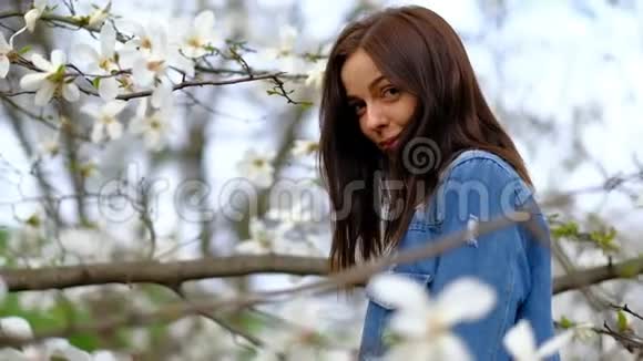 年轻迷人的苗条女子穿着蓝色时髦的牛仔裤夹克摆在盛开的木兰树和鲜花上视频的预览图