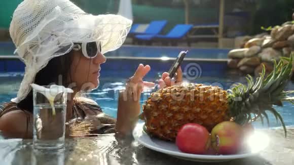 年轻漂亮的女人在游泳池里用手机在盘子旁边喝水吃热带水果3840x2160视频的预览图