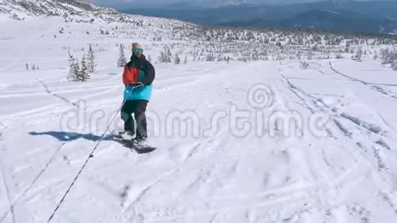 在一个阳光明媚的冬天在雪山上骑着一条绳子穿着颜色鲜艳的夹克和太阳镜的快乐男子滑雪板视频的预览图