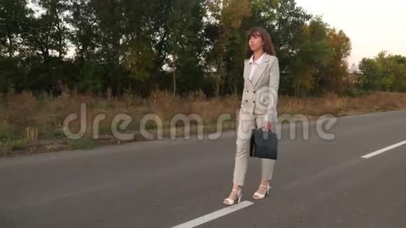 女商人带着一个黑色公文包穿着浅色西装穿着白色高跟鞋沿着沥青路走上面有标记视频的预览图