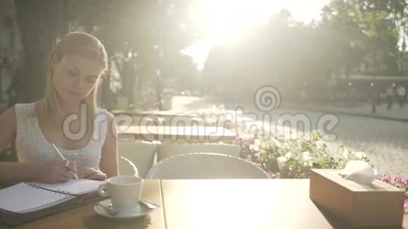 身着白衣的金发年轻女性在咖啡馆外的露台桌上写着个人日记日记中的思绪眺望视频的预览图
