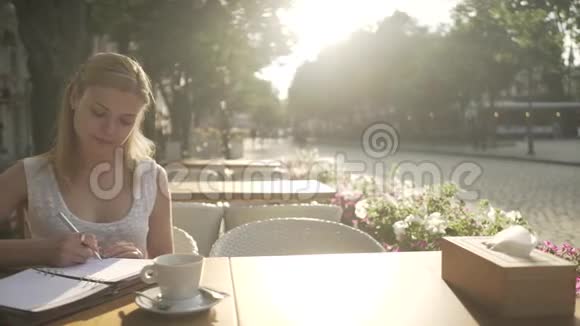 穿着白色裙子的金发少女在咖啡馆外的露台桌上写着个人日记日记看着远方视频的预览图