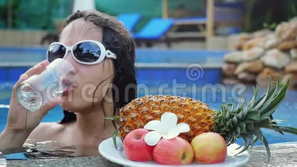 年轻女子戴着墨镜一边喝水一边在游泳池里慢慢地晒着水果和鸡蛋花视频的预览图