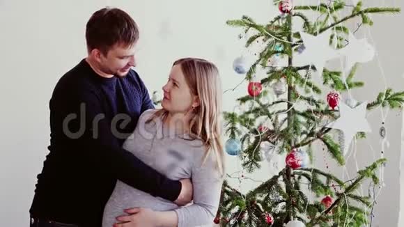 幸福的年轻夫妇靠近美丽的圣诞树丈夫拥抱他怀孕的妻子慢动作3840x2160视频的预览图