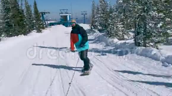 在一个阳光明媚的冬天在雪山上骑着一条绳子穿着颜色鲜艳的夹克和太阳镜的快乐男子滑雪板视频的预览图