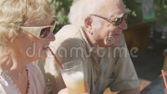 快乐的老年夫妇戴着太阳镜坐在户外咖啡馆里喝着鸡尾酒视频的预览图
