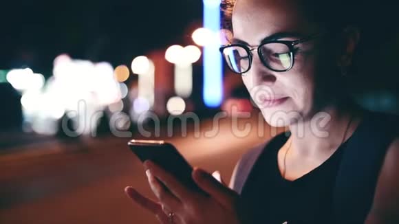 在一个夜晚的城市里漂亮的女人一边用手机在街上行走在后台可以看到视频的预览图