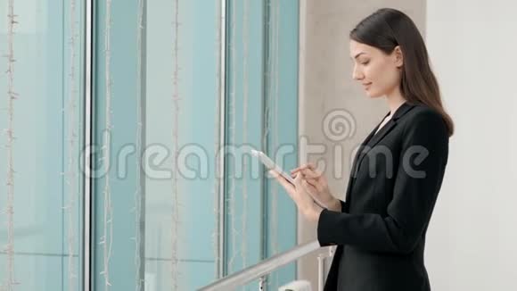 漂亮的女商人在办公室大楼的窗户前站着一边用数码平板电脑俯瞰视频的预览图
