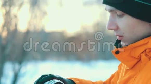 一个年轻人在冬天的公园里喝热水瓶里的热饮料大雪纷飞穿着橙色夹克和温暖衣服的人视频的预览图