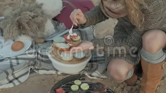 穿着外套的年轻女子坐在河边海边的海滩上野餐在烤架上煮肉和蔬菜视频的预览图