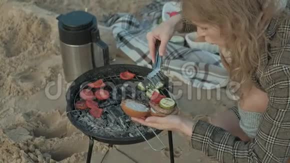 穿着外套的年轻女子坐在河边海边的海滩上野餐在烤架上煮肉和蔬菜视频的预览图
