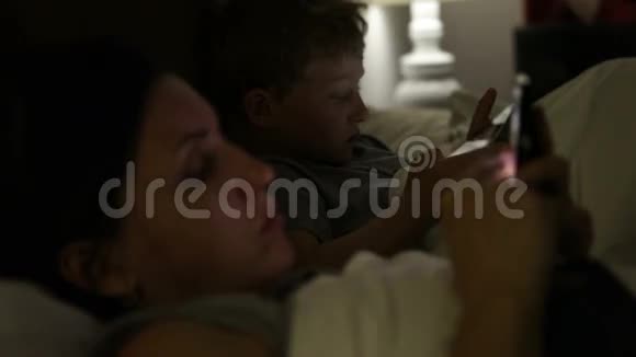 母亲和儿子躺在床上看着他们的电子设备视频的预览图