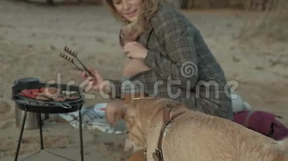 一位穿着外套的年轻女子坐在河边海边的海滩上野餐在烤架上煮肉和蔬菜视频的预览图
