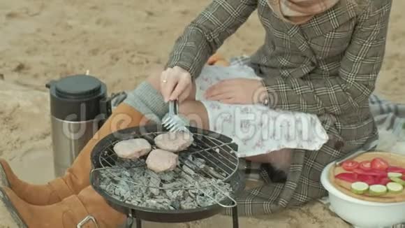 穿着外套的年轻女子坐在河边的海滩上大海野餐在烤架上煮肉寒冷的天气视频的预览图