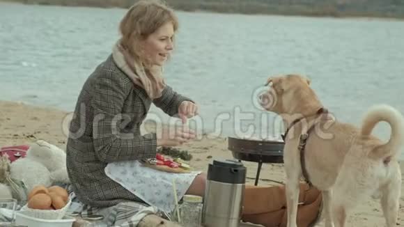一位穿着外套的年轻女子坐在河边海边的海滩上野餐在烤架上煮蔬菜和肉一个视频的预览图