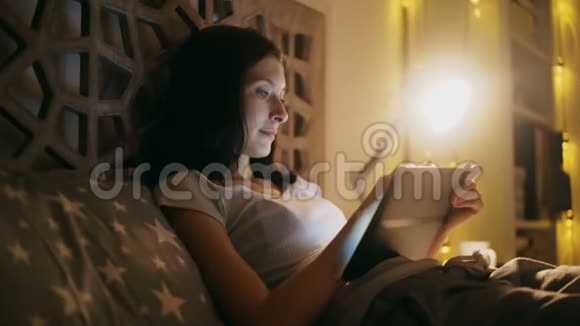年轻的笑脸女人晚上躺在家里的床上用平板电脑上网购物视频的预览图