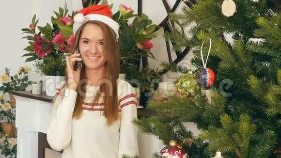 戴着红圣诞帽的微笑女孩在圣诞树附近的电话里交谈视频的预览图
