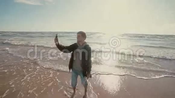 一个年轻人光着脚站在海滩上牛仔裤卷起试图自拍视频的预览图