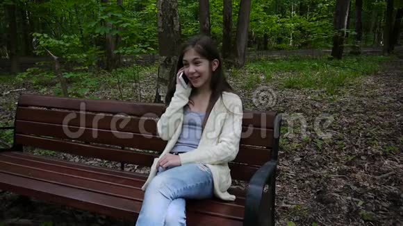 年轻迷人的女孩在长凳上和电话交谈夏日公园微笑高清视频镜头拍摄静态摄像机视频的预览图