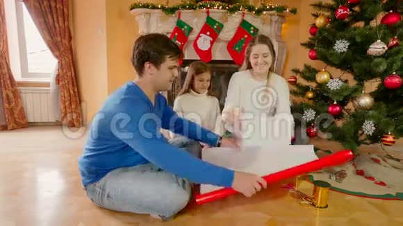 年轻的幸福家庭坐在客厅里用纸包圣诞礼物视频的预览图