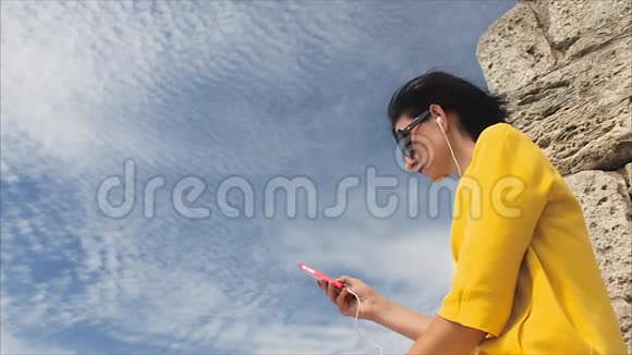 有魅力的女人在天空和古代废墟的背景下在智能手机上用耳机听音乐视频的预览图