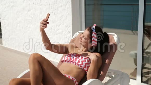 微笑的女人躺在躺椅上在智能手机上自拍视频的预览图