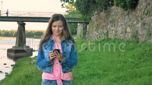 漂亮的年轻女孩用智能手机沿着海滨散步背景中的野石桥和老墙视频的预览图