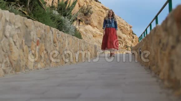 穿红色裙子的浪漫女孩走在海边的路上视频的预览图