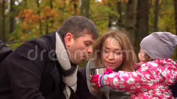 可爱的小女孩和她的父母在秋天公园里从真空瓶里喝着热气腾腾的茶家庭野餐快乐4K视频的预览图