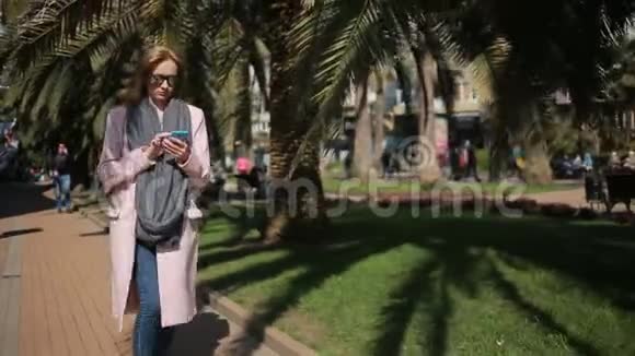 年轻女子微笑着在公园里用手机聊天沿着小巷和棕榈树散步视频的预览图