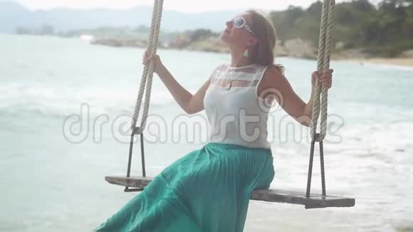 漂亮的年轻快乐的女人戴着太阳镜在石头海滩的秋千上缓慢地放松1920x1080视频的预览图