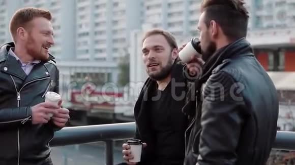 三个英俊的年轻人带着胡子说话微笑着喝咖啡在城市的一座桥栏杆附近慢节奏视频的预览图