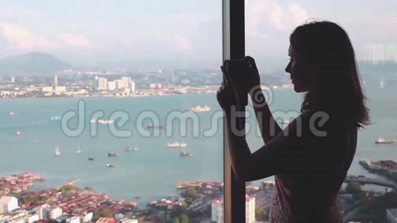 一位年轻的女游客站在窗前在手机上看到槟城摩天大楼的高度视频的预览图