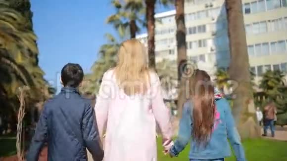 一个女人带着孩子一个母亲牵着双胞胎的手漫步在一条有棕榈树的大道上视频的预览图