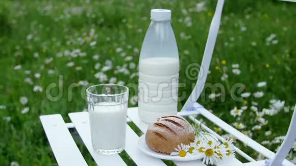 在洋甘菊草坪的中间白色的椅子上有一瓶牛奶还有一杯牛奶和面包附近视频的预览图