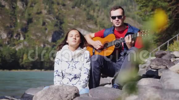 海滩上的一对夫妇在山河旁边的夏天弹吉他唱歌1920x1080视频的预览图